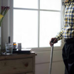 Elderly man standing with a cane next to a table with flowers and medication