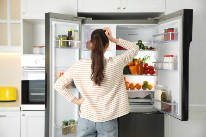 A woman standing in front of an open refrigerator filled with various fruits and vegetables