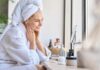 Dermatologists’ Secret to Softer Skin Revealed Woman in a bathrobe with a towel on her head, smiling at her reflection in a mirror