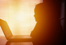 Silhouette of a person working on a laptop with sunlight streaming through blinds