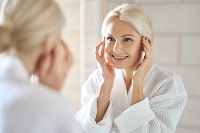shutterstock_2142010179.jpg Smiling woman in a bathrobe applying skincare in front of a mirror