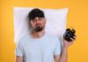 Man wearing a sleep mask holding an alarm clock with a frustrated expression