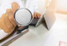 A doctor holding a stethoscope in an office setting with a laptop in the background