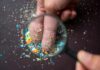 Hidden Pollutant Dwarfs PFAS Exposure A hand holding a magnifying glass over fingers covered in colorful particles