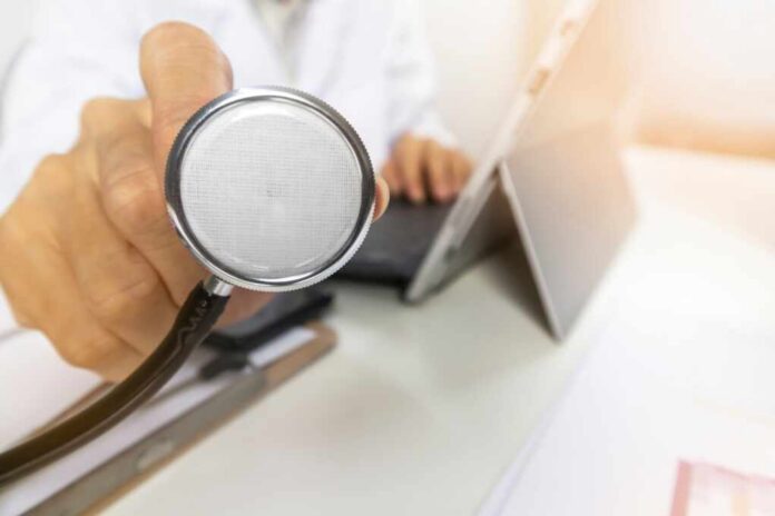 A doctor holding a stethoscope in an office setting with a laptop in the background