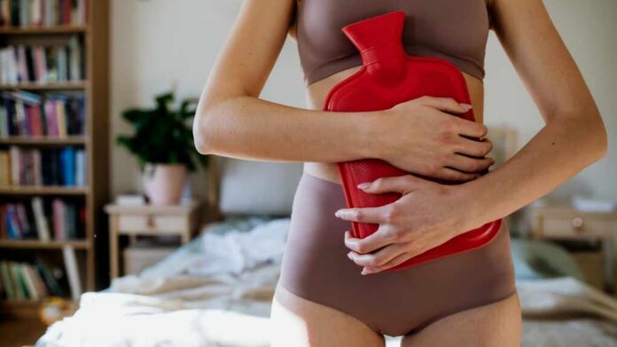 Woman in a cozy bedroom holding a red hot water bottle against her abdomen