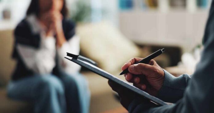 Writing,,Clipboard,And,Counseling,With,Hands,Of,Person,For,Psychologist, A therapist taking notes during a session with a client in the background