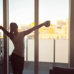 Woman stretching with coffee by a balcony window at sunrise