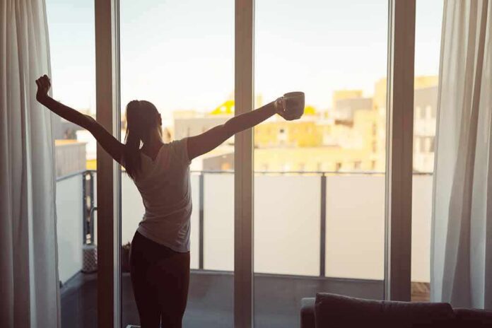 shutterstock_1016532013 Woman stretching with coffee by a balcony window at sunrise