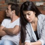woman looking down while man sits with arms crossed on couch