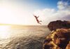 Person jumping off a rocky cliff into the ocean at sunset