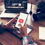 Hands holding smartphone with video app icon at a workspace