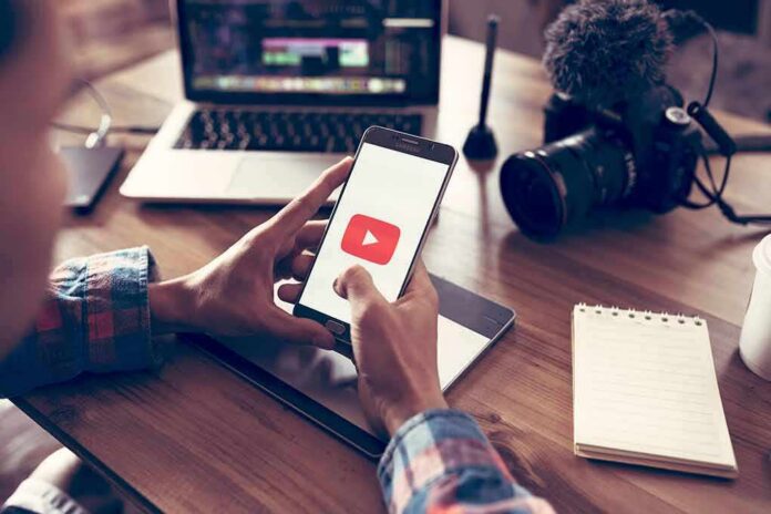 shutterstock_1041026248 Hands holding smartphone with video app icon at a workspace