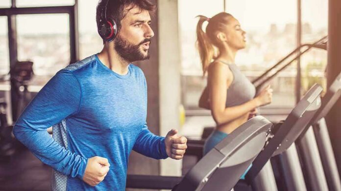 shutterstock_1256975917 Man in blue long-sleeve running on a treadmill with headphones