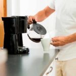 Person pouring coffee from carafe into mug beside drip coffee maker