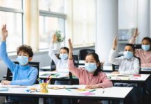 Schoolchildren wearing masks raise hands in a classroom