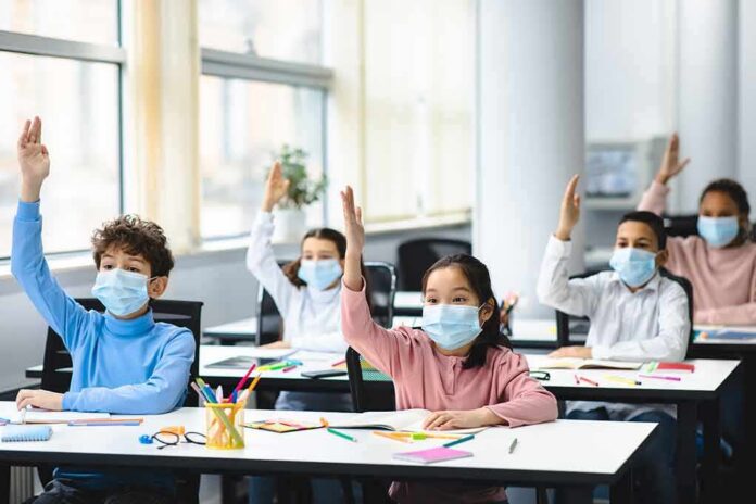 Schoolchildren wearing masks raise hands in a classroom
