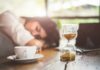 Vitamin Red Flag Tied To Crushing Fatigue Hourglass on table with coffee cup and sleeping person in background