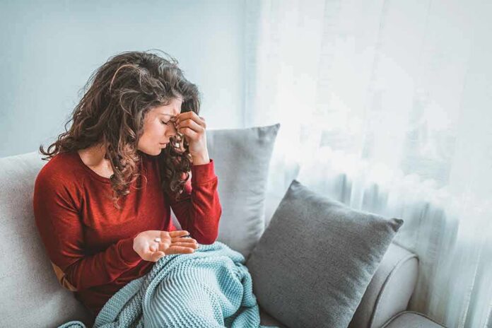 shutterstock_1508066993-1 Woman sitting on couch holding pills and rubbing her forehead
