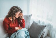 Midlife Migraine Spike: What’s Really Driving It Woman sitting on couch holding pills and rubbing her forehead