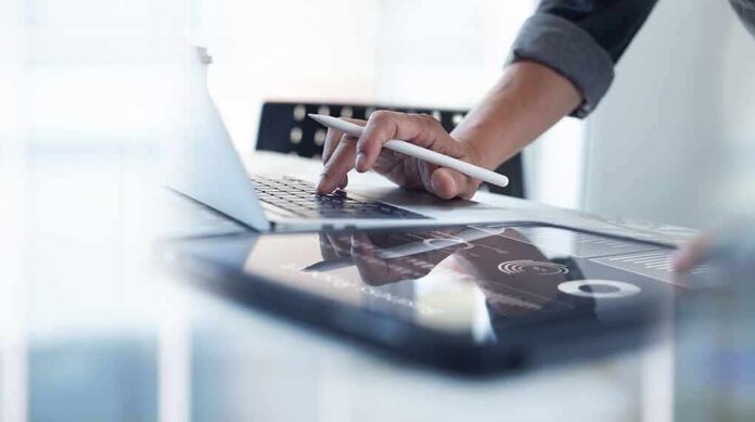 shutterstock_2147248723 Person using laptop with stylus near tablet